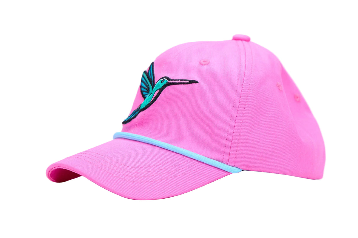 A bright pink baseball cap featuring an embroidered hummingbird logo in teal and black on the front, with a light blue accent stripe along the edge of the curved brim, set against a plain white studio background.
