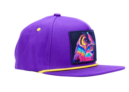 A purple flat-brim baseball cap with a yellow contrast brim edge and a colorful embroidered owl patch on the front, set against a plain white background.