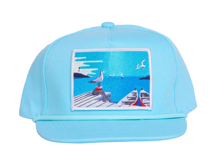 Aqua blue flat bill hat with a patch on the front featuring an ocean pier with a seagull and a boat
