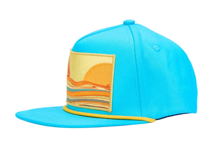 A bright turquoise blue flat-brim cap with a rectangular embroidered patch on the front depicting a yellow and orange sun over layered stripes, finished with a yellow trim along the edge of the brim.