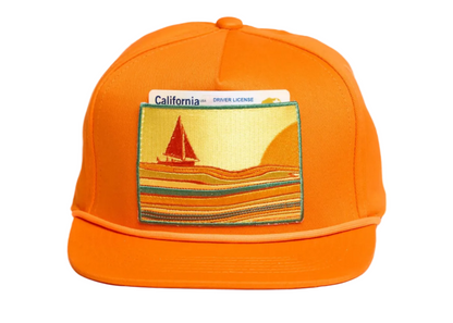 Bright orange snapback cap featuring a rectangular embroidered patch on the front with a sailboat scene and a California driver license card tucked behind it.