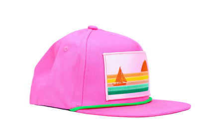 A bright pink baseball cap with a flat brim, featuring a colorful embroidered patch on the side and a thin green accent line along the edge of the brim, set against a light blue background.