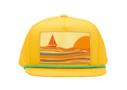 A bright yellow flat-brim cap featuring a rectangular embroidered patch on the front with a sailboat and sunset scene in orange, yellow, and teal stripes, set against a plain white background.