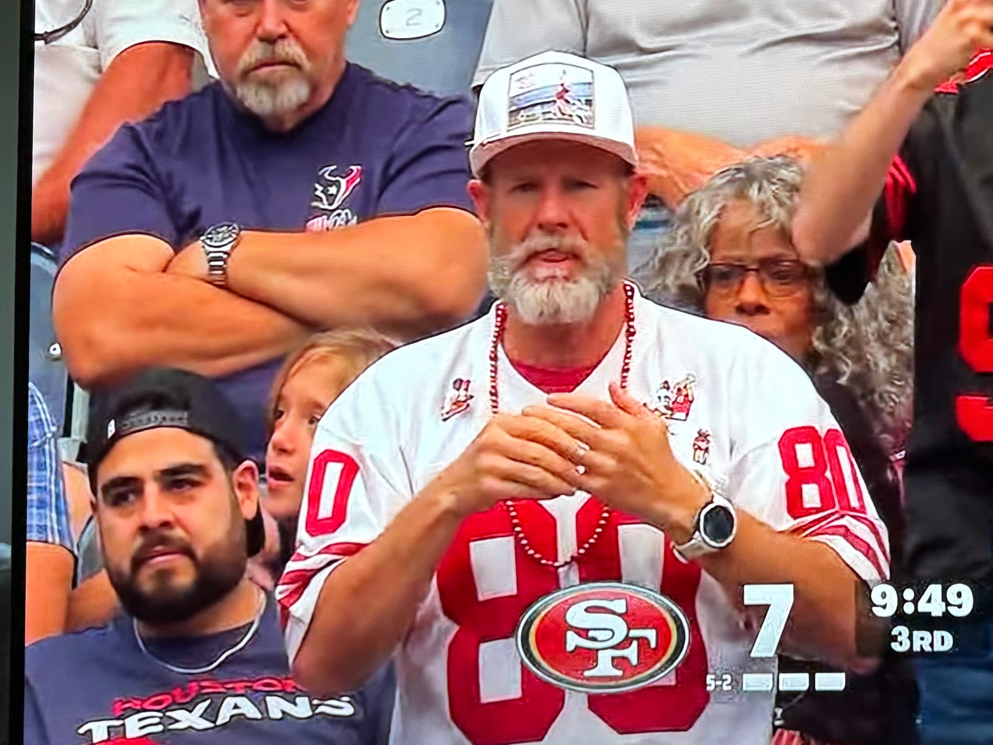 An image taken from a TV broadcast of a 49ers fan wearing a white hat featuring a photographic patch with an image of The Catch, the legendary touchdown caught by Dwight Clark of the 49ers
